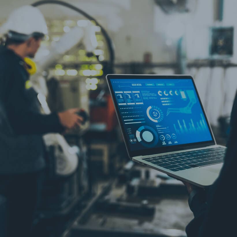 A man in a hardhat holds a laptop in front of a large industrial machine, focused on his work.